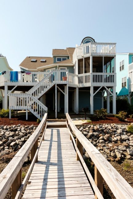 Stilted blue waterfront beach house with multi-level decks and a wooden walkway over stone riprap.