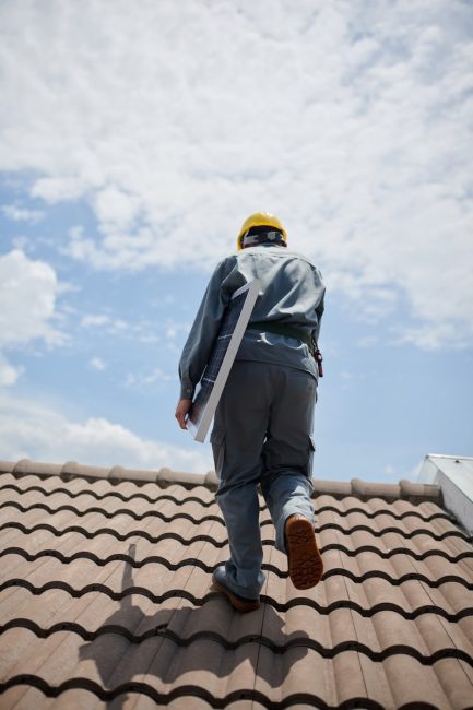 A roofing professional wearing a yellow hard hat walks on a tiled roof while carrying a solar panel, showcasing the services of a fortified roof expert in Hattiesburg, MS.