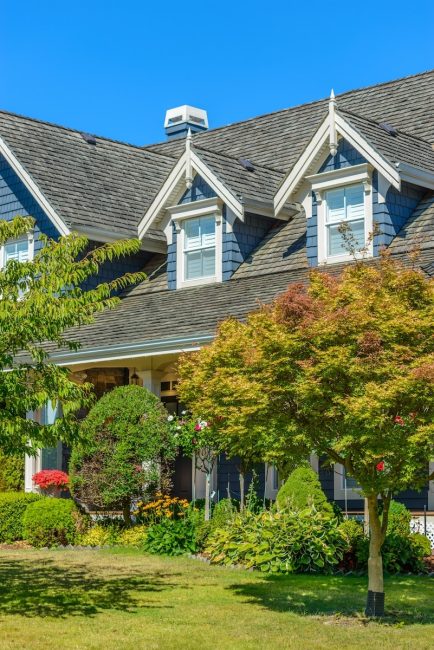 Suburban home with blue siding, prominent dormer windows, and a landscaped front yard under a clear blue sky.